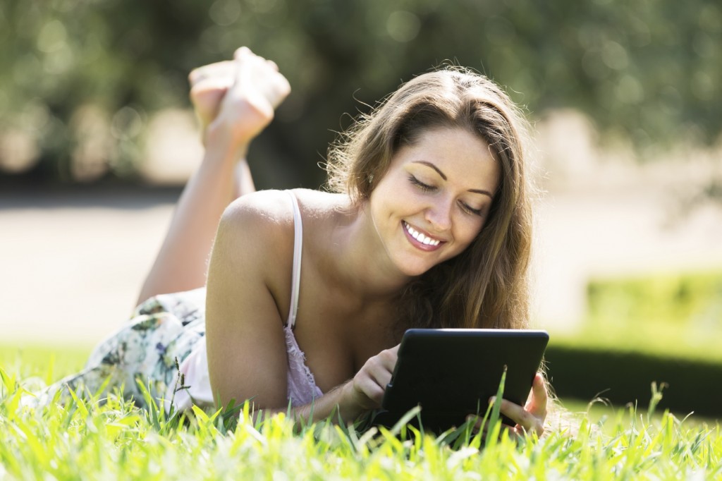 Attractive smiling girl lying on grass in meadow and using tablet