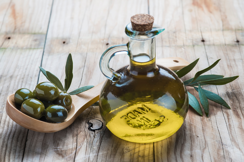 A glass bottle of olive oil and a wooden spoon with  olives on a table